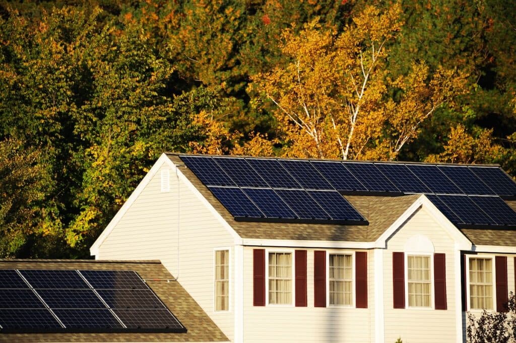 A house with solar panels on the roof, a proud example of solar installation in Middleton, is surrounded by trees boasting rich autumn foliage. Sunlight illuminates the panels, accentuating the vibrant green and yellow leaves in the background.
