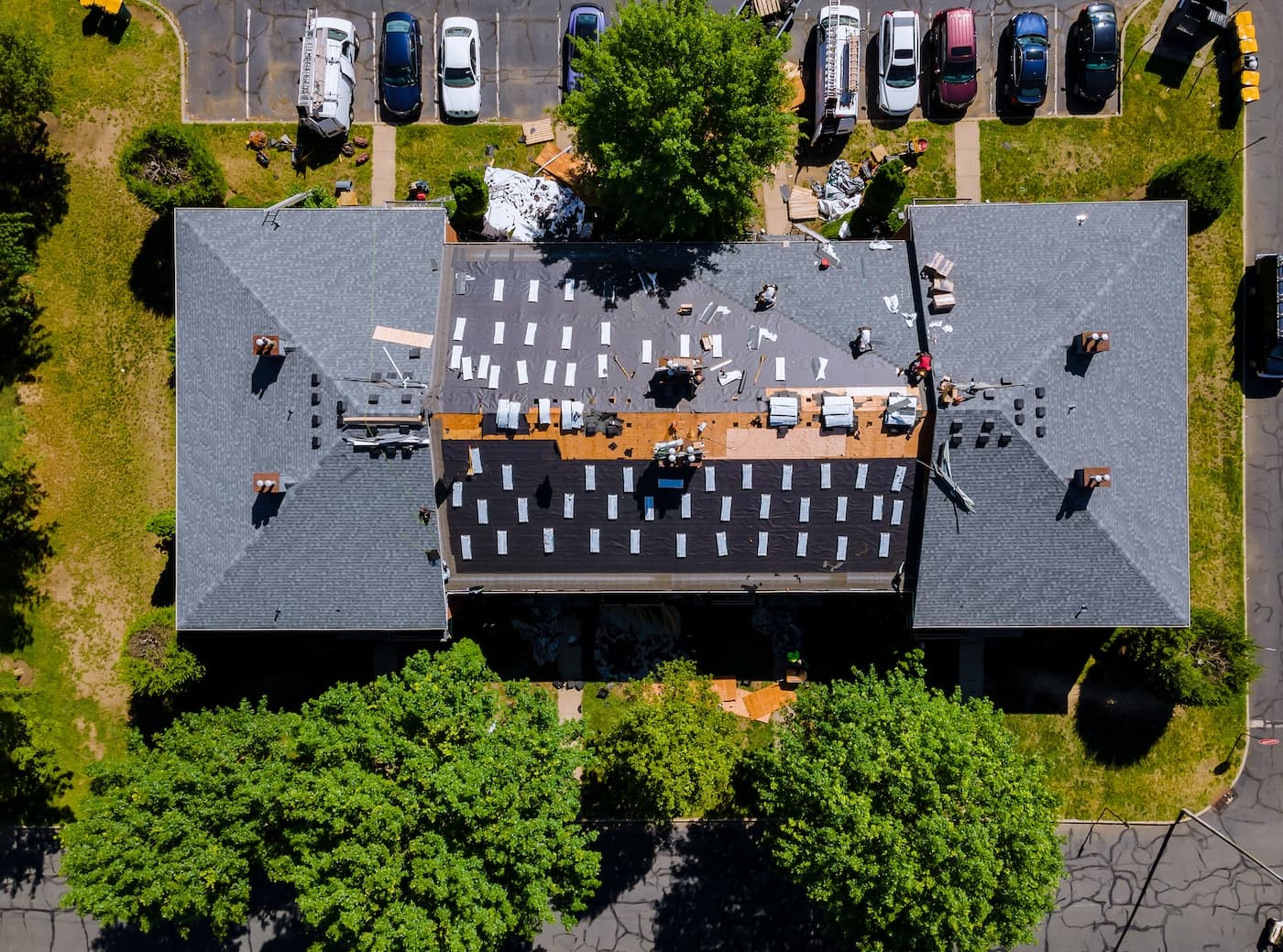 Aerial view of a building with workers replacing the roof; shingles and materials are scattered on the rooftop, and cars are parked nearby. Trees surround the building, and construction equipment is visible.