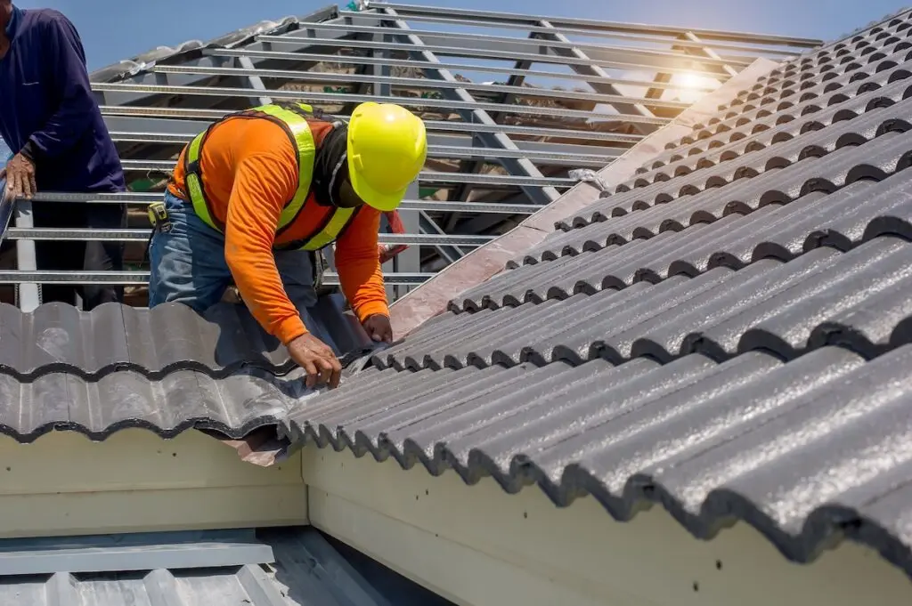 A construction worker in a yellow helmet and safety vest installs gray roof tiles on a house, while another stands nearby—demonstrating how long does it take to replace a roof under sunny skies.