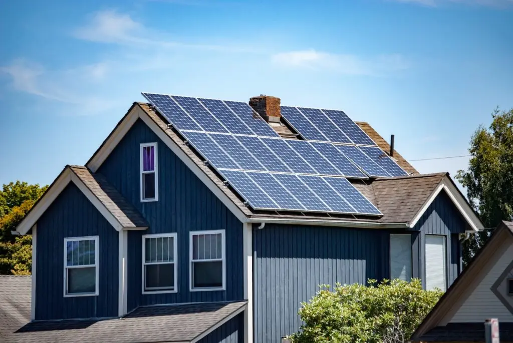 A blue house with white trim has multiple solar panels installed on its roof, under a clear blue sky, surrounded by trees and neighboring homes.