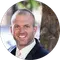 A man in a suit and tie is smiling with a blurred outdoor background featuring a tree and some greenery.