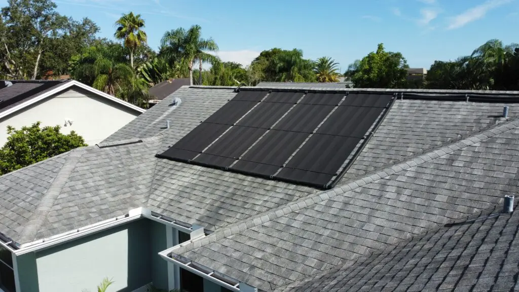 A rooftop with gray shingles features several black solar panels installed on one section, offering a glimpse of how many solar panels to power a house. Surrounding homes, trees, and a clear blue sky are visible in the background. how many solar panels to power a house