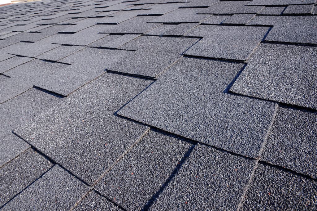 Close-up view of overlapping gray asphalt roof shingles arranged in a uniform pattern, highlighting their texture and areas of granular loss on the roof.