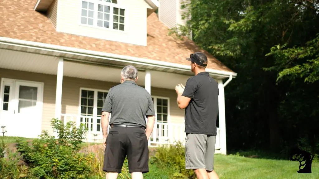 Two men stand outside, facing a beige house with large windows and a porch, surrounded by greenery. The sun casts light on the scene, highlighting the relaxed, summery atmosphere.