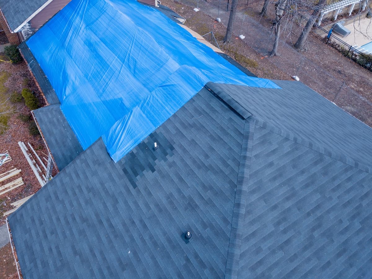 Aerial view of a house with a roof partially covered by a large blue tarp. The exposed sections of the roof display dark gray shingles. Surrounding the house are some bare trees and construction materials on the ground.