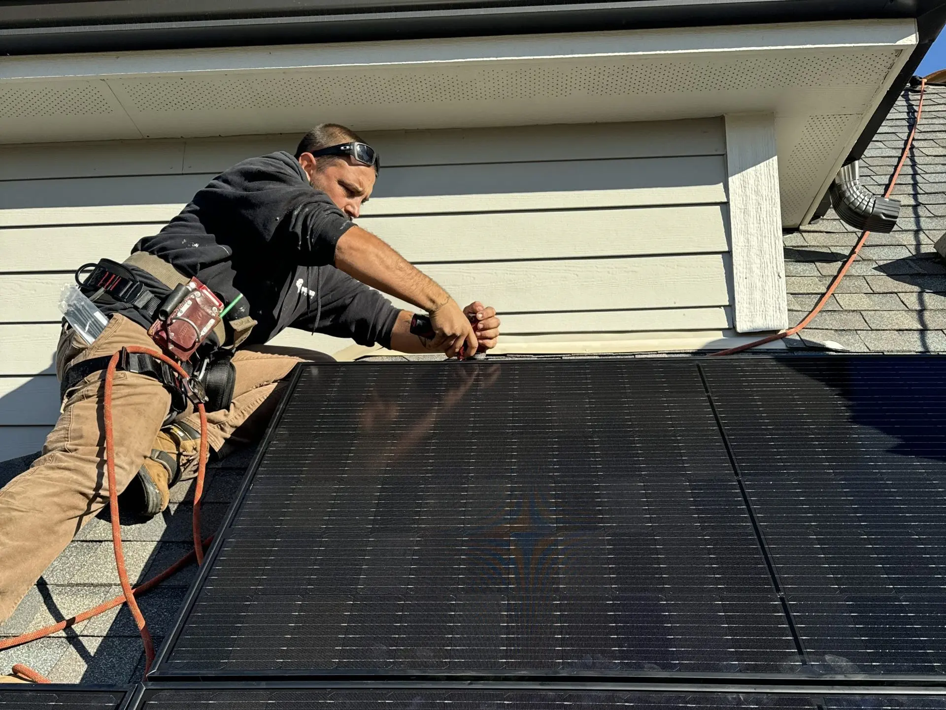 A worker in safety gear installs a large black solar panel on a rooftop, using tools and harnesses, on a sunny day.