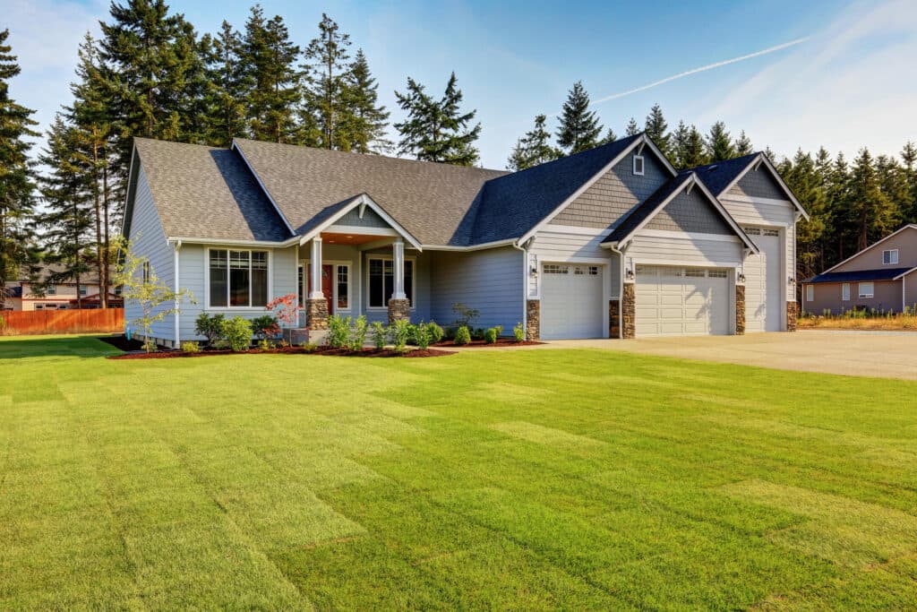 A modern single-story house with a gray and white exterior, a neatly manicured lawn, a covered front porch, and a double garage. Tall trees and a clear sky provide a serene background.