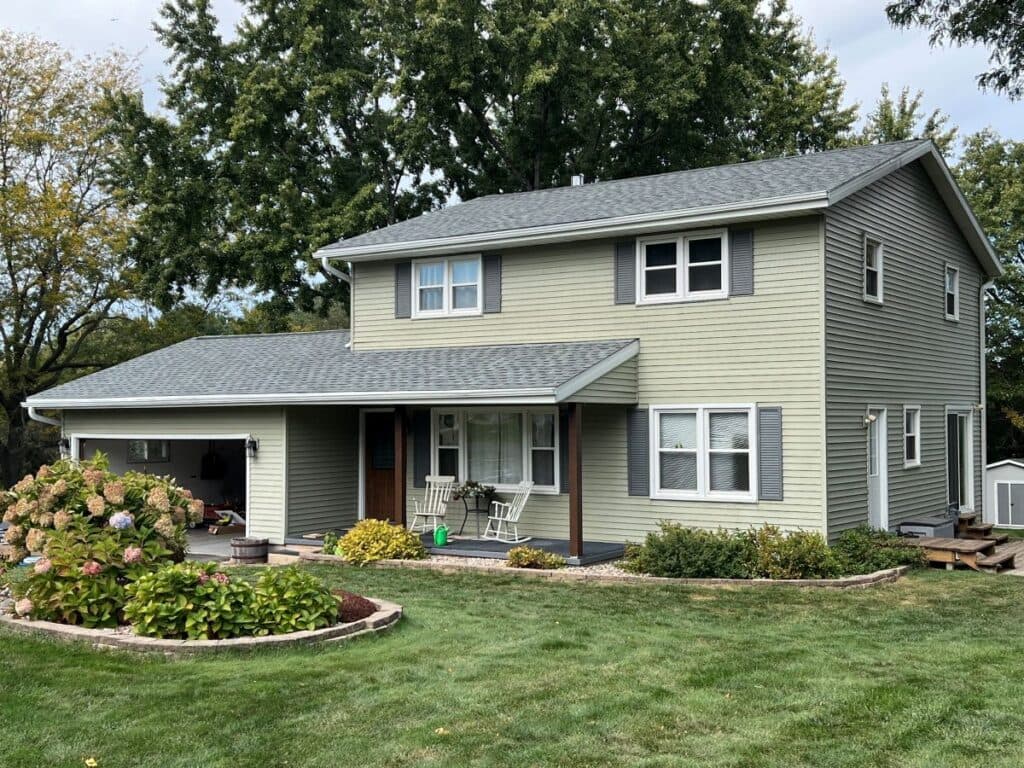 A two-story light green house with gray shingles, white trim, and an attached garage. The front lawn is neatly mowed with bushes and flowers near the entrance, and large trees are visible in the background.
