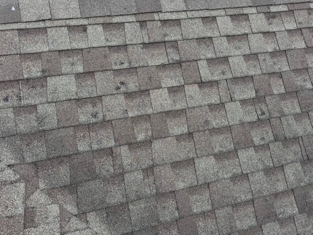 Close-up view of an asphalt shingle roof with a mix of light and dark brown rectangular shingles arranged in an overlapping pattern; some shingles show minor dark spots and weathering.