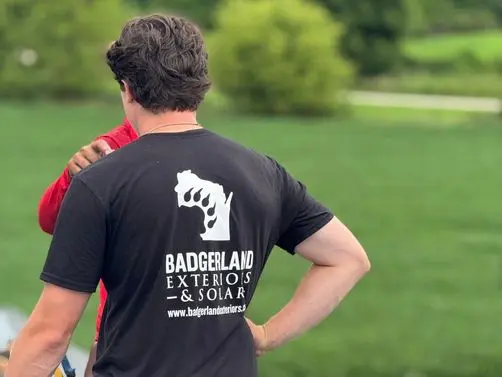 roofing companies waunakee wi workers on top of shingles roof A person in a “Badgerland Exteriors & Solar” t-shirt stands on a roof with another person, near roofing materials, with green grass and trees in the background.