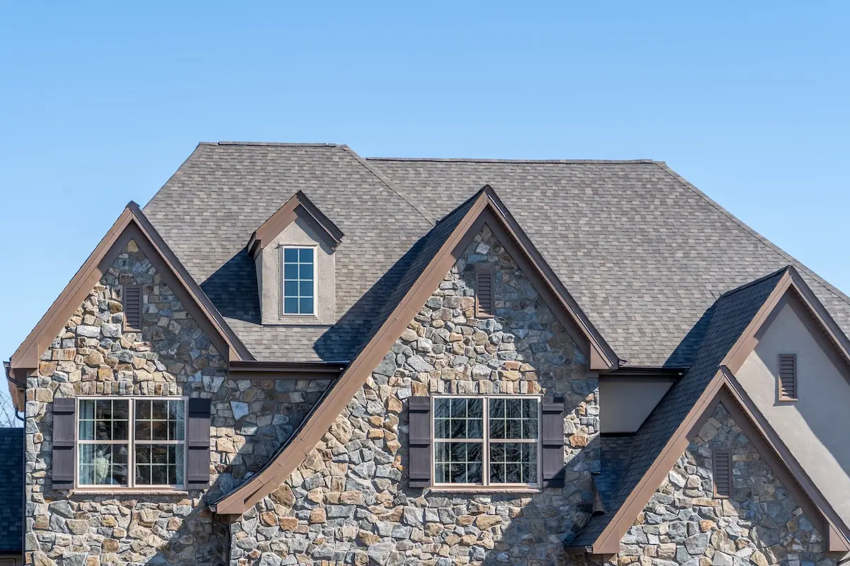 The image shows the upper portion of a house with stone and tan siding, featuring multiple peaked roofs, dormer windows, and brown trim under a clear blue sky.