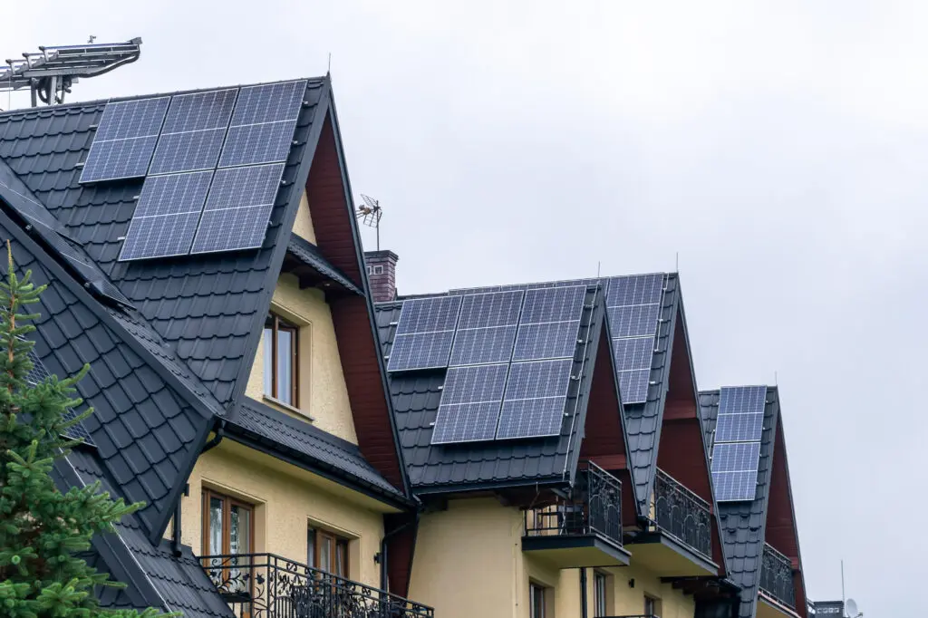Row of houses with dark, sloped metal roofs, each fitted with solar panels on metal roof. The buildings have balconies with ornate railings, and the sky is overcast.