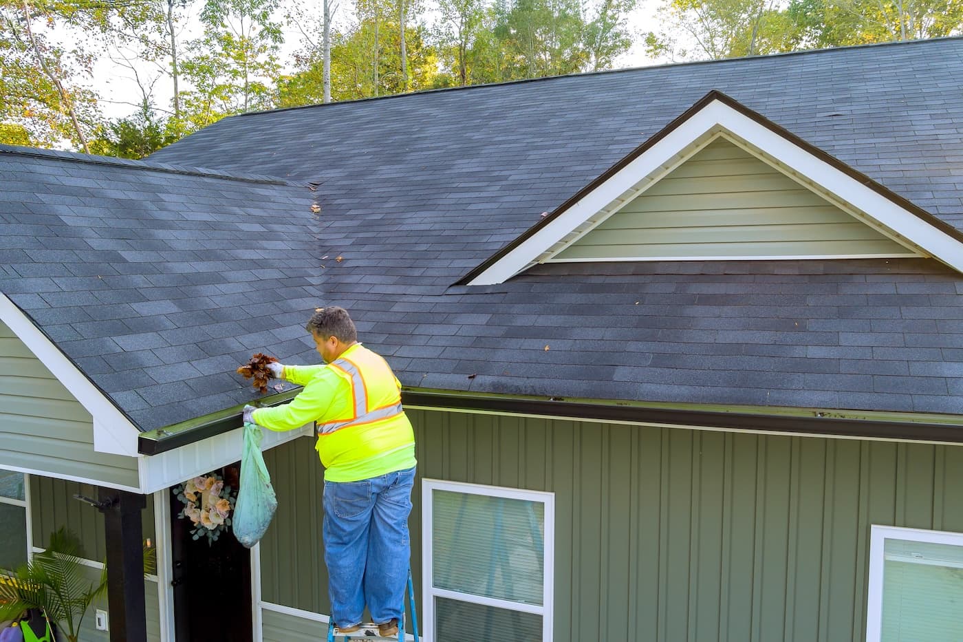 A person in a neon safety vest stands on a ladder, cleaning leaves and debris from the gutter of a green house with a sloped roof. Trees are visible in the background.
