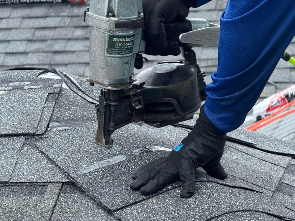roofing companies waunakee wi workers on top of shingles roof A person wearing gloves uses a nail gun to install asphalt shingles on a roof, securing overlapping gray shingles in place.