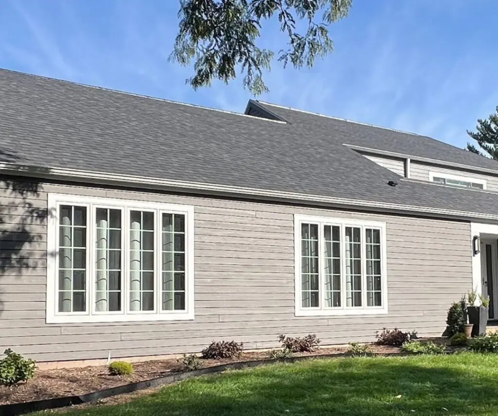 A gray house with a dark gray roof by roofing New Berlin, featuring white-trimmed tall windows and a small garden in front, set against a blue sky with tree branches at the top.