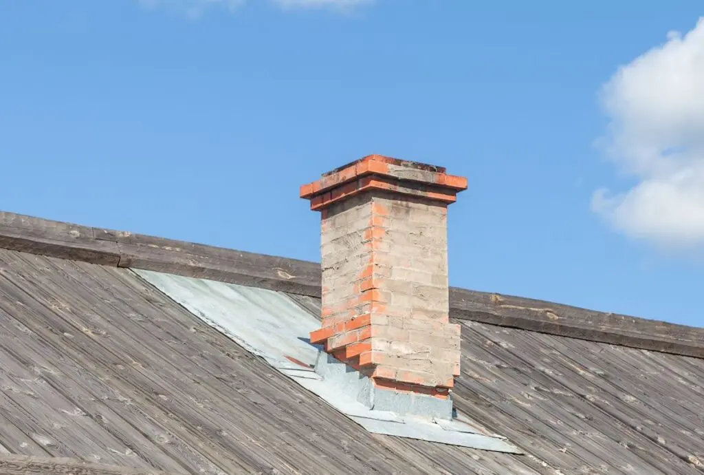 A brick chimney extends from a slanted wooden roof, with metal flashing around its base. The sky is clear and blue with a few white clouds.