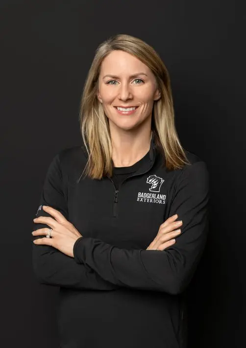 A woman with straight blond hair smiles, standing with arms crossed. She wears a black zip-up pullover with the logo and text 
