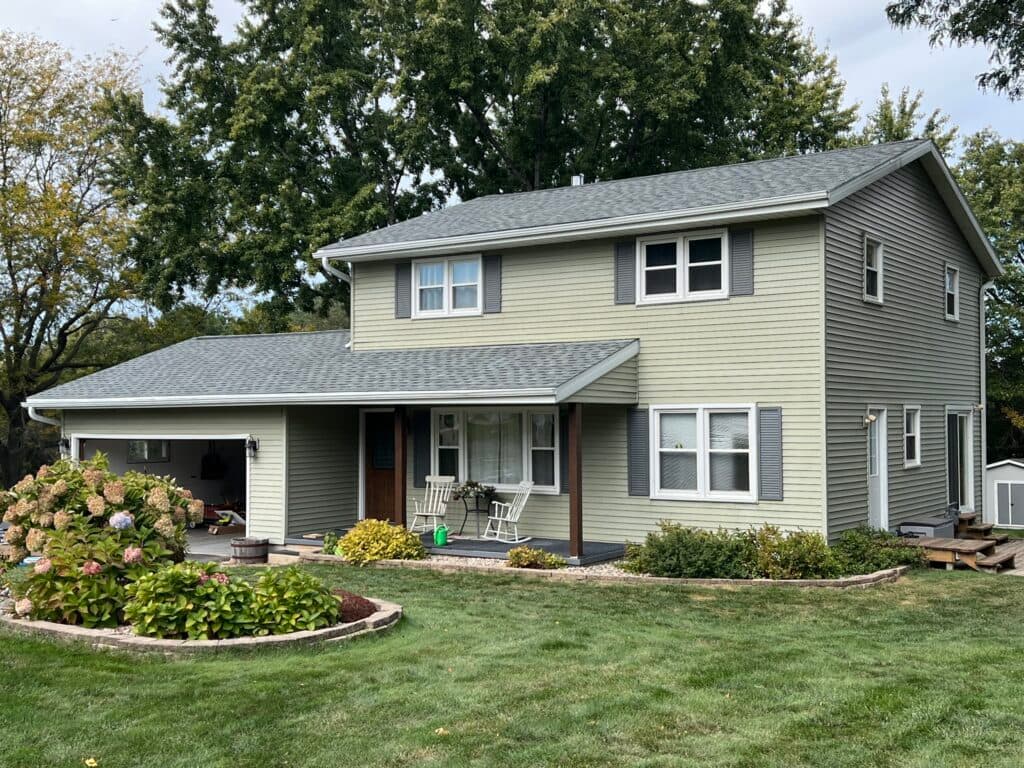 asphalt shingle roof replacement A two-story light gray house with white trim, a covered porch, attached garage, and well-maintained yard with green grass, bushes, and blooming hydrangeas in front.