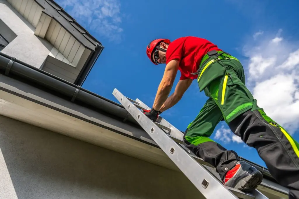 A worker in safety gear and a red helmet stands on a ladder, cleaning or inspecting the gutter of a house under a blue sky with clouds.