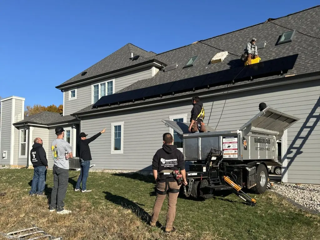 Six workers install solar panels on the roof of a gray house using equipment and a trailer on a sunny day. A ladder lies in the grass in the foreground.