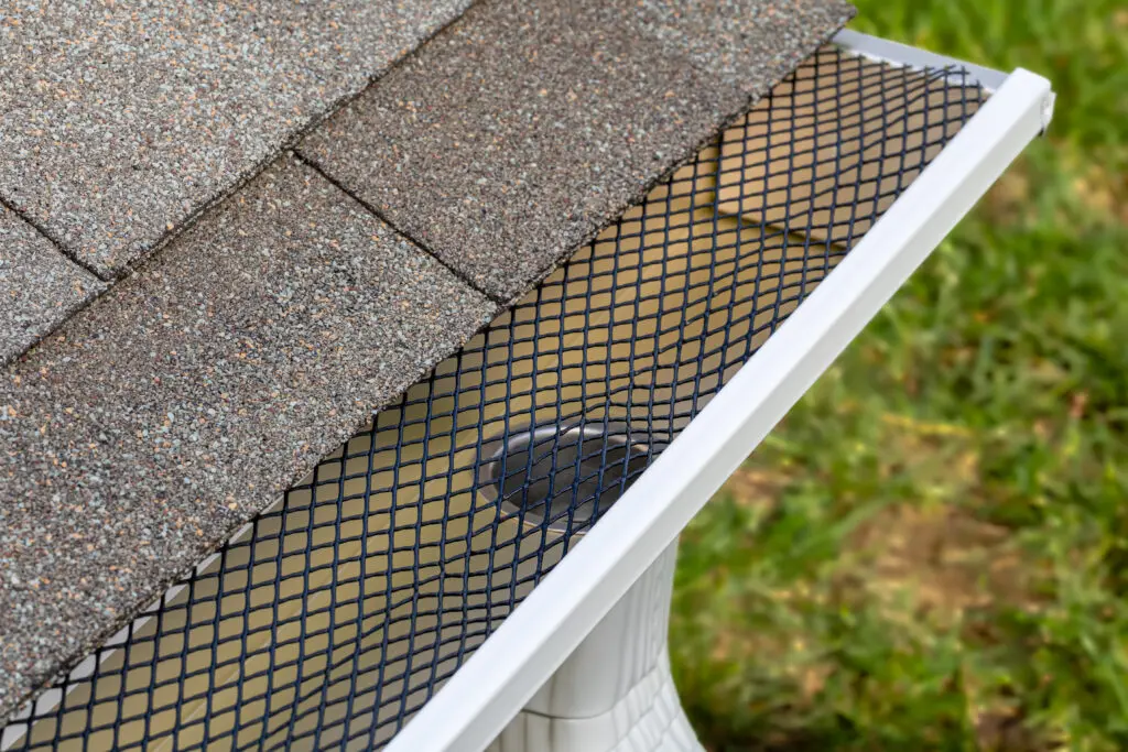 A close-up view of a house roof with asphalt shingles and a gutter covered by a black mesh guard, prompting the question: are gutter guards worth it for keeping debris out? Green grass is visible in the background. are gutter guards worth it