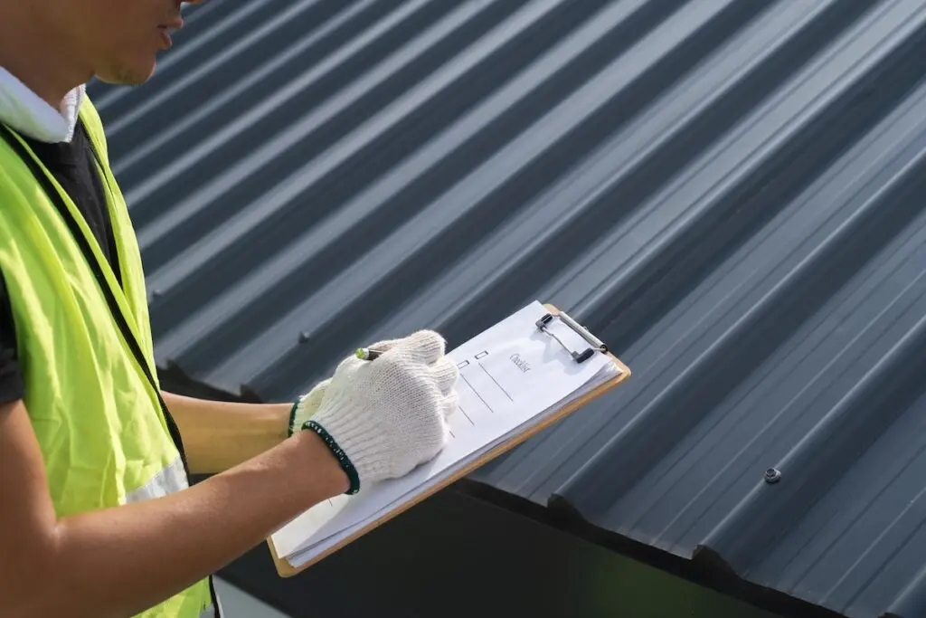 A person wearing a safety vest and white gloves holds a clipboard and pen, conducting a commercial roof inspection next to a corrugated metal roof. Only part of their face and upper body is visible.