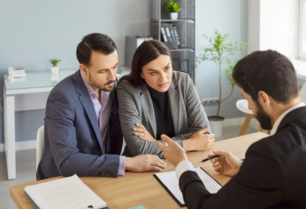 hail damage roof repair  man and woman sit closely together at a desk, attentively listening to a professional in a suit who is explaining documents and gesturing with a pen in an office setting.