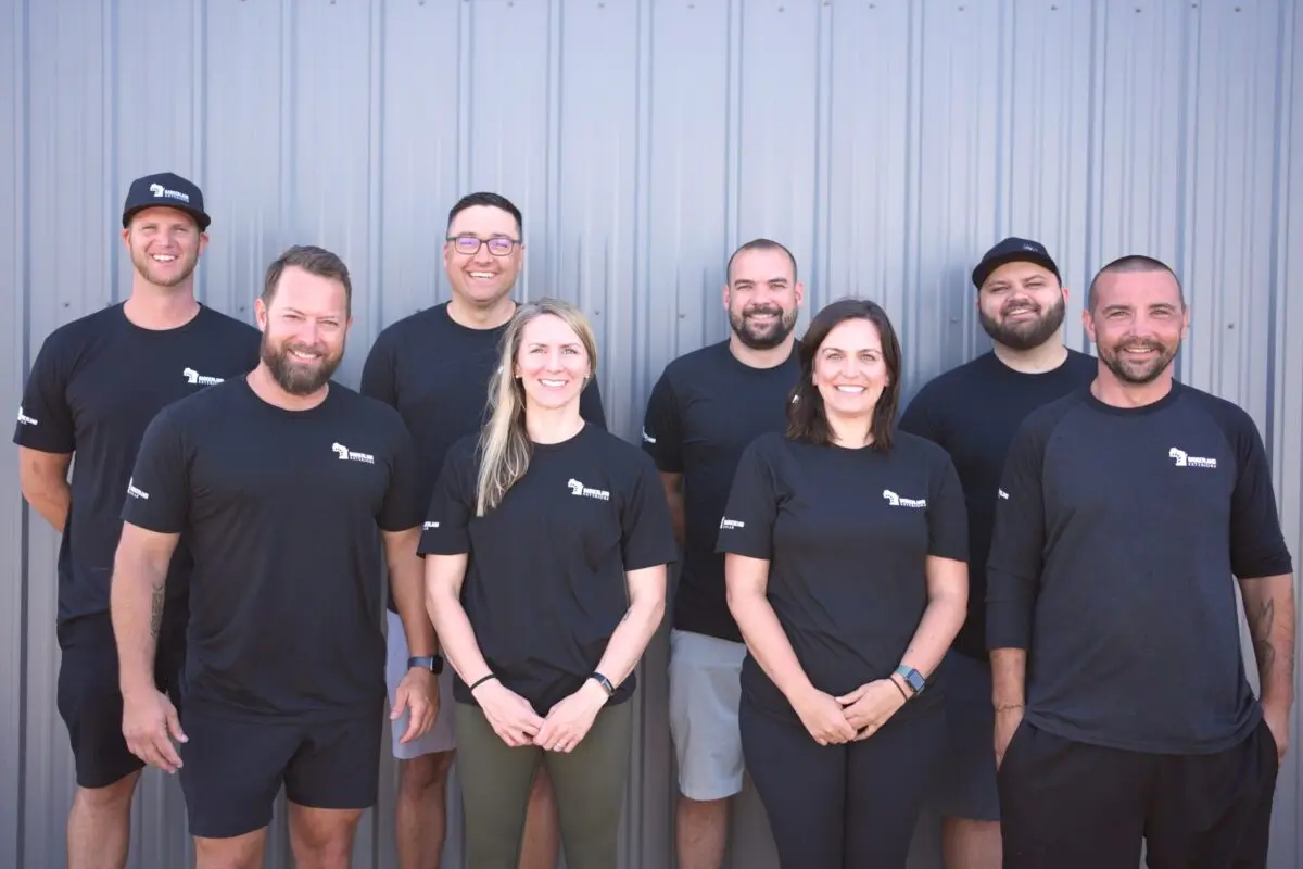 roofing companies verona wi Eight people stand in two rows, smiling in front of a corrugated metal wall. They all wear matching black shirts with a small white logo on the chest. The group appears friendly and casual.