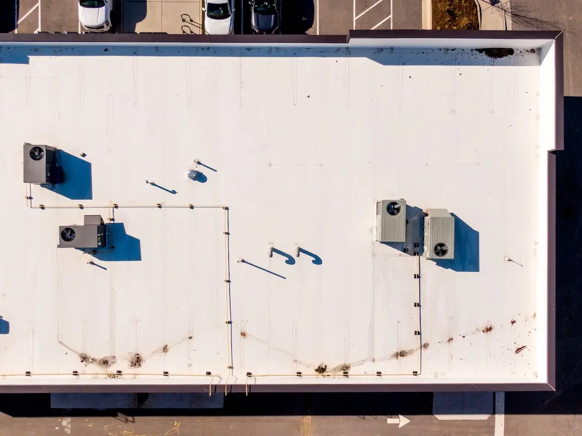 Aerial view of a flat white rooftop, possibly TPO vs EPDM, with several HVAC units and vents. Some dirt and debris are scattered near the edges. Parking spaces and parked cars are visible along the perimeter of the building.