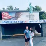 A man and a woman stand in front of a building decorated with a large American flag, eagle, and Statue of Liberty replica. The words "Land of the Free N' Home of the Brave" are displayed above them. Trees are in the background.