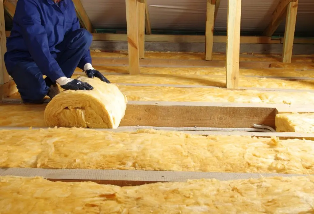 A person in blue work clothes and gloves is unrolling yellow insulation material in an attic with wooden beams and a sloped roof.