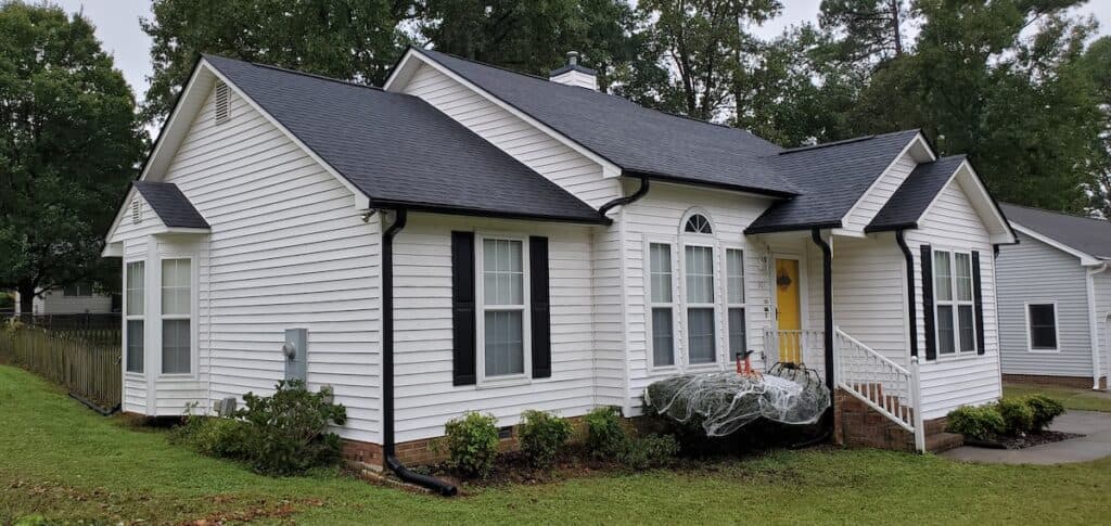A white single-story house with black shutters and a dark roof, featuring shrubs along the front, a small porch with a railing, and Halloween decorations on the bushes. Trees and a lawn are in the background.