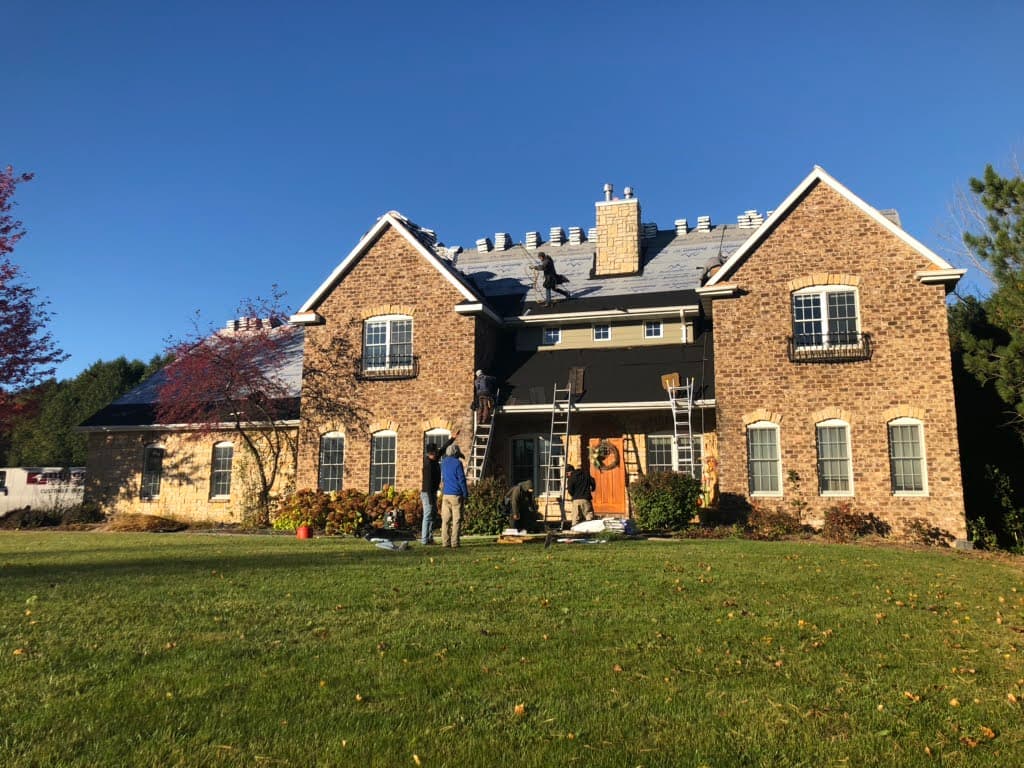 Workers are installing or repairing the roof of a large two-story brick house on a clear, sunny day. Ladders lean against the house and tools are scattered on the grass in the foreground.