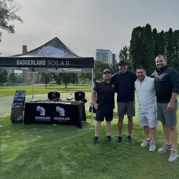 Four men stand together on a golf course near a Badgerland Solar tent with promotional materials. Trees and buildings are visible in the background.