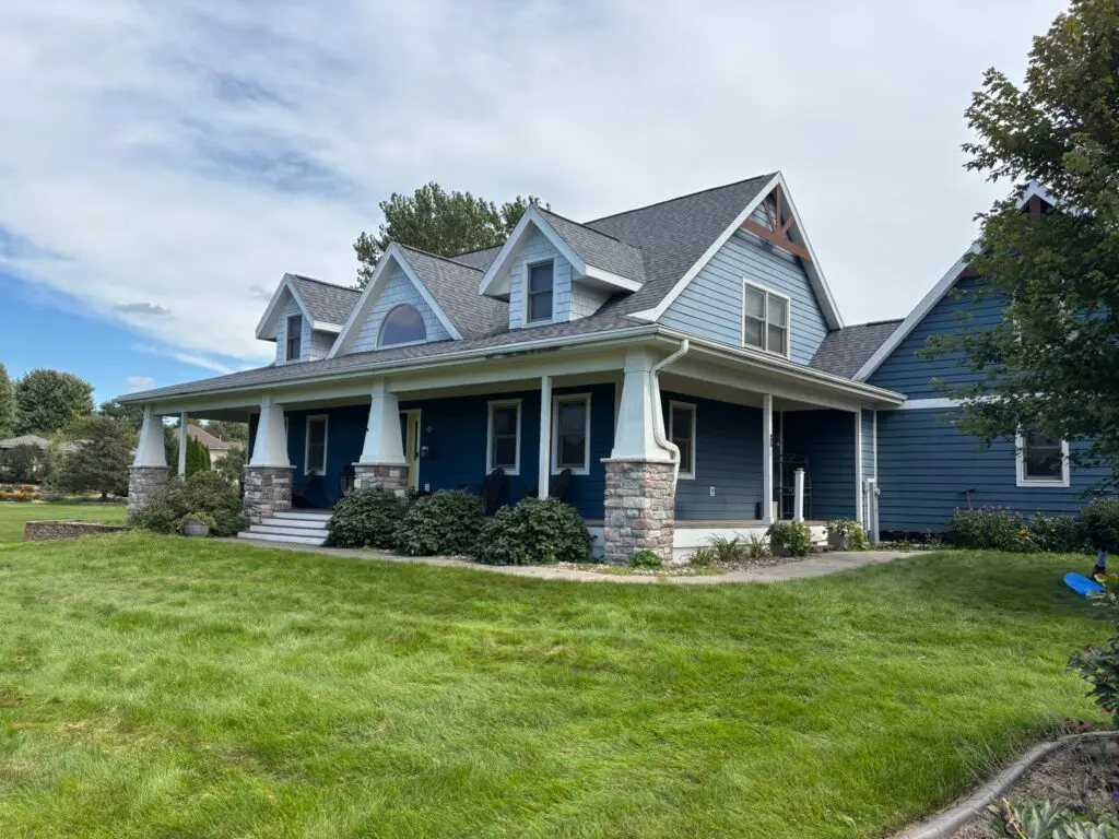 A two-story blue house with white trim, large front porch with columns, and stone accents sits on a well-kept green lawn under a partly cloudy sky.