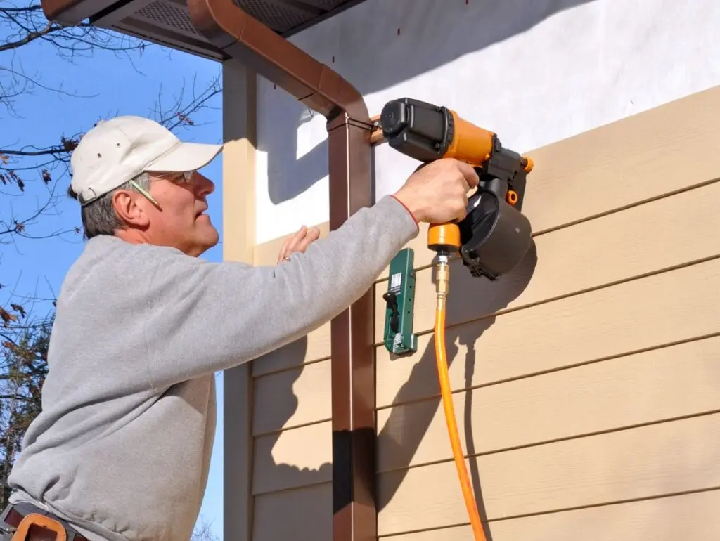 fiber cement siding installation A person standing on a blue ladder uses a nail gun to install tan siding on the exterior wall of a house on a sunny day. The person is wearing a gray sweater, beige cap, and a tool belt.