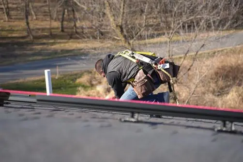 roof maintenance checklist A worker wearing a safety harness bends over while working on a roof, with trees and a road in the blurred background.