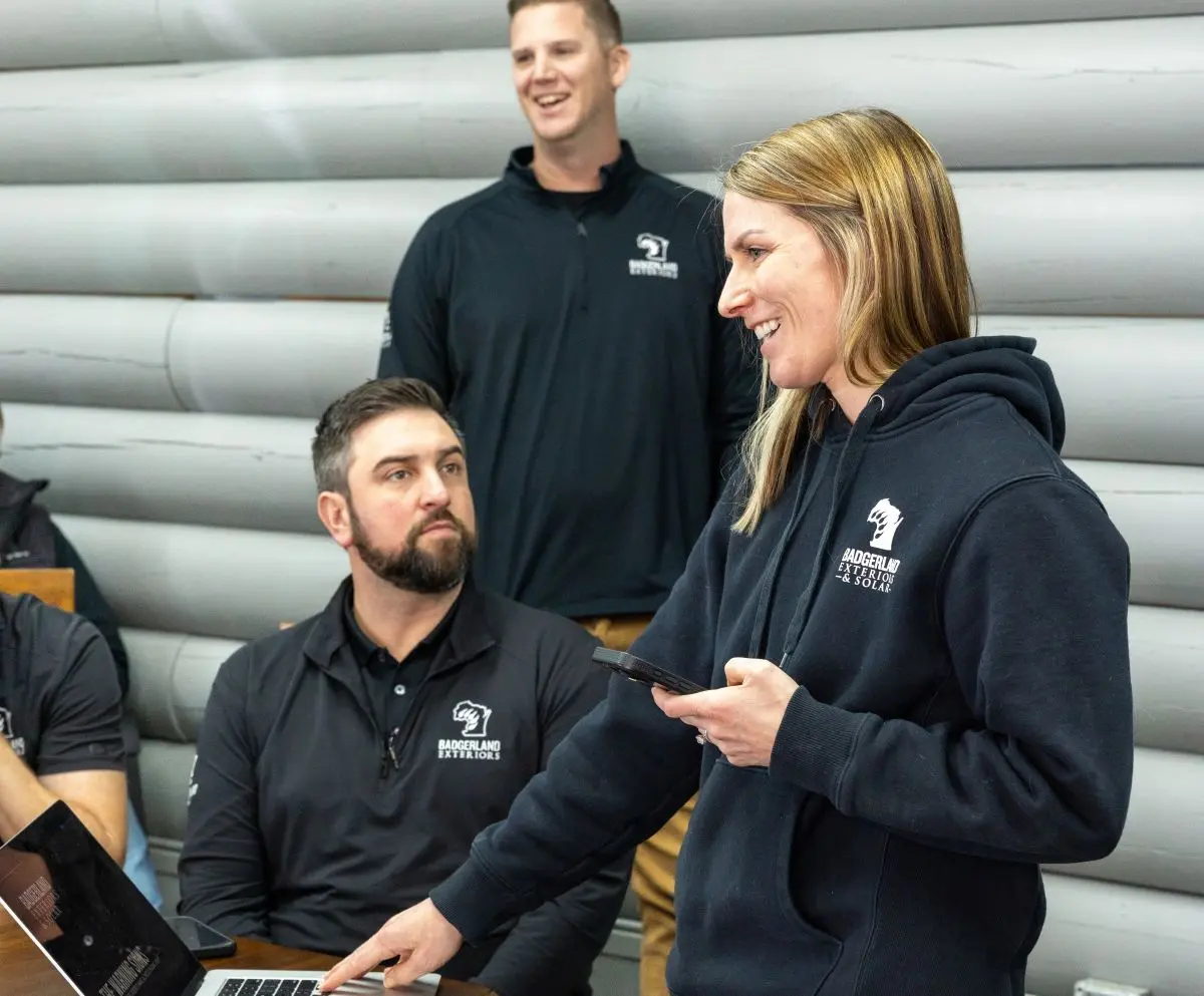 A woman in a hoodie smiles while holding a phone and pointing at a laptop, standing with three colleagues in front of a gray wooden wall. Two men sit at a table, and one man stands smiling in the background.