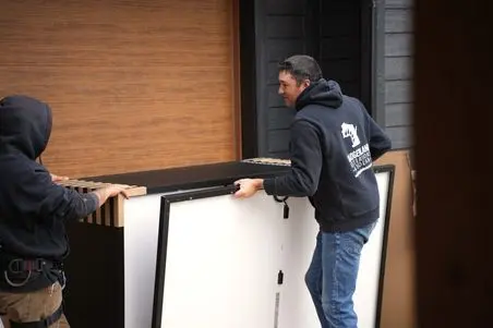 Two people wearing hoodies are carrying a large rectangular panel together in front of a wooden garage door. Both are focused on moving the item carefully.