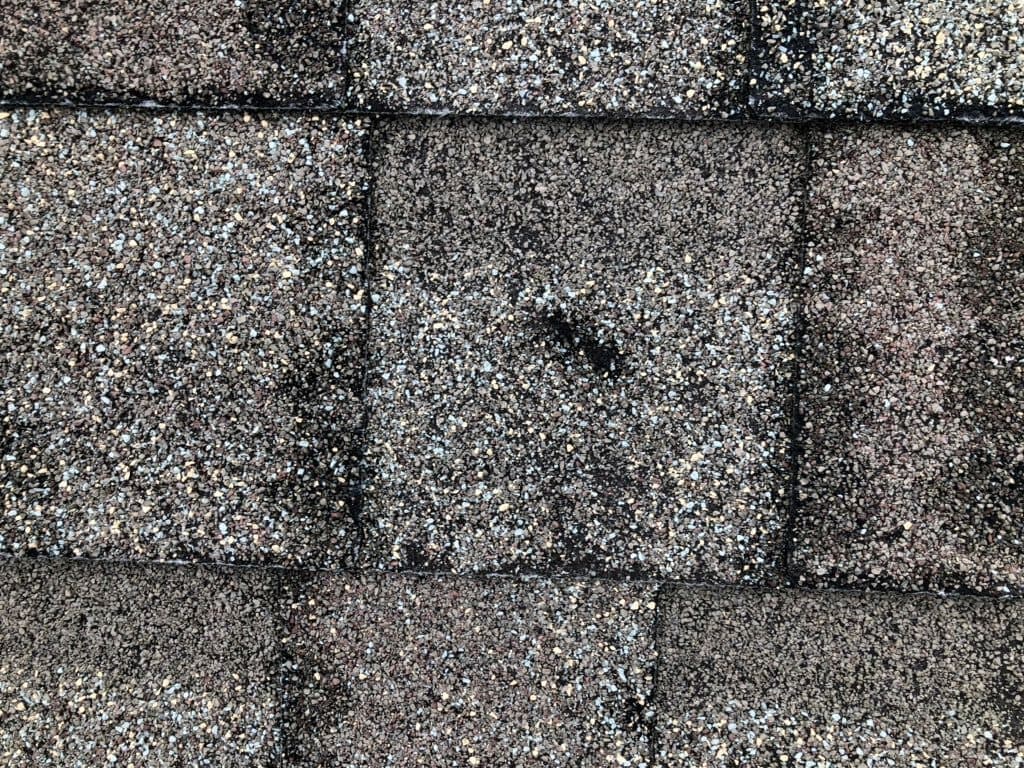 Close-up view of weathered asphalt roof shingles showing a mix of brown, gray, and black granules, with some visible wear and darkened spots indicating age or possible water damage.