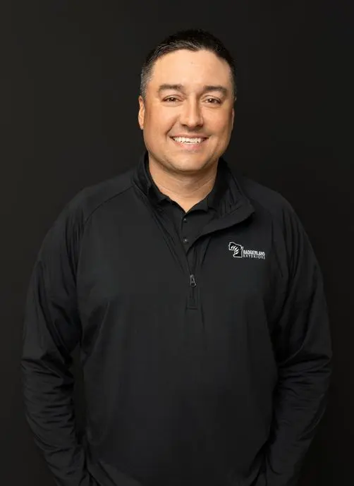 A man with short dark hair smiles at the camera, wearing a black zip-up jacket with an embroidered logo reading 
