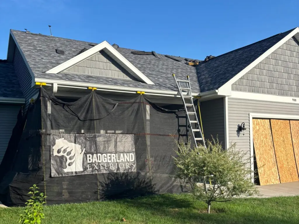 roofing companies verona wi A house with gray siding and boarded-up garage has roofing work underway. A ladder leans against the roof, and a black protective covering with "Badgerland Exteriors" shields the front porch area.