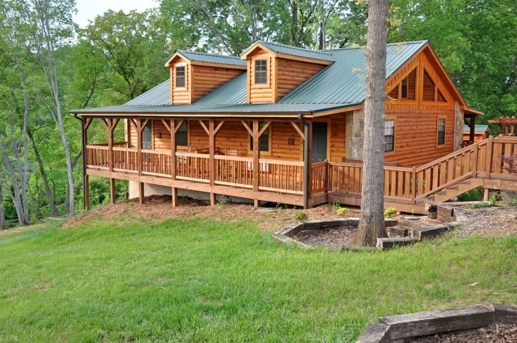A wooden log cabin with a green metal roof and wraparound porch sits on a grassy hillside surrounded by trees and lush greenery. Steps lead up to the cabin’s entrance.