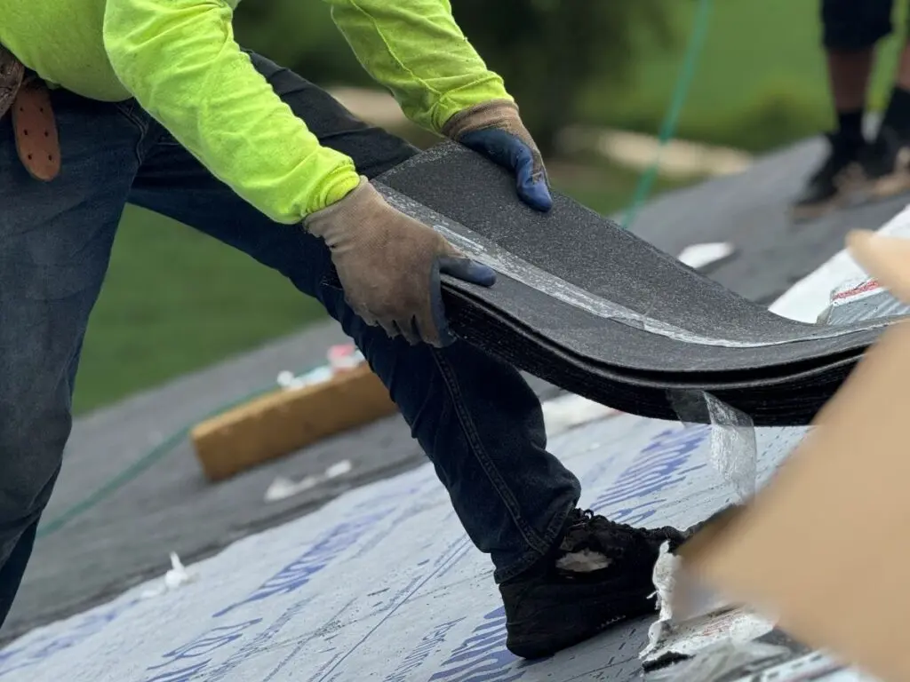 wind damage roof repair worker in a neon yellow shirt and gloves installs shingles on a roof, with tools and roofing materials scattered nearby. The worker is bent over, placing a shingle on roofing underlayment.