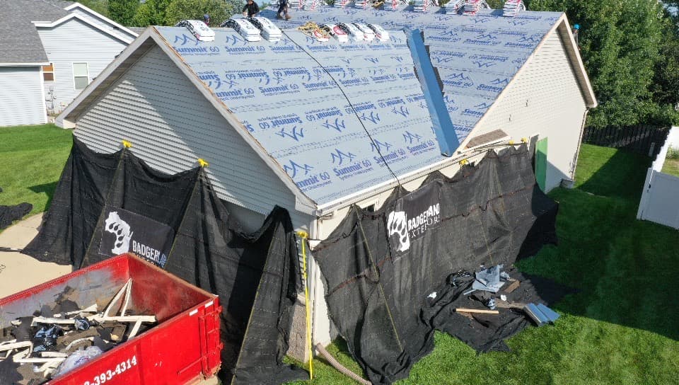 A house with its roof under construction, covered in underlayment and roofing materials. Black tarps protect the sides, debris is scattered below, and a red dumpster holds old roofing material.