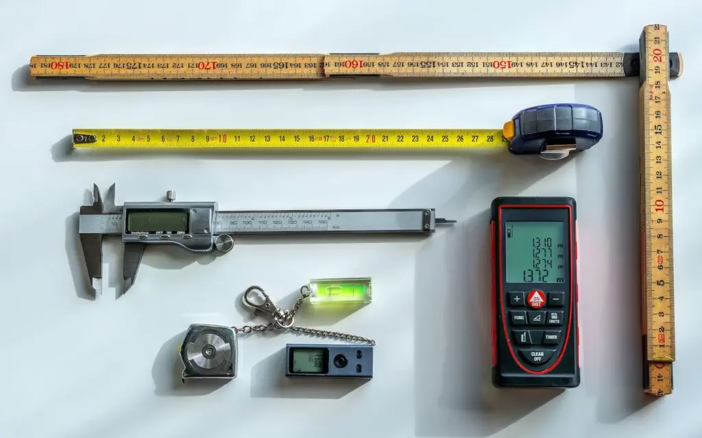 A variety of measuring tools are arranged on a white surface, including tape measures, a ruler, a digital caliper, a laser distance meter, a plumb bob, and a small digital level.