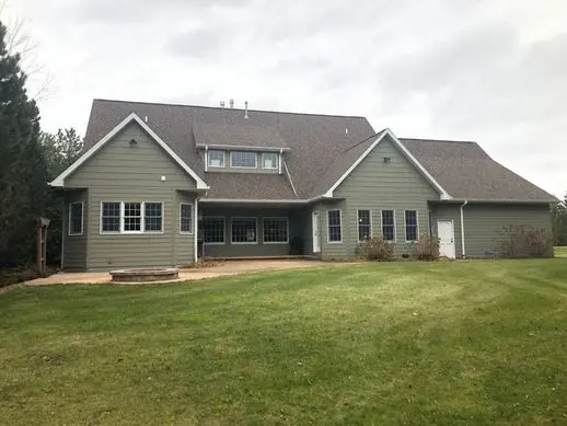 A large two-story house with green siding and a brown shingle roof. The yard is grassy with a small patio and surrounded by trees under a cloudy sky.