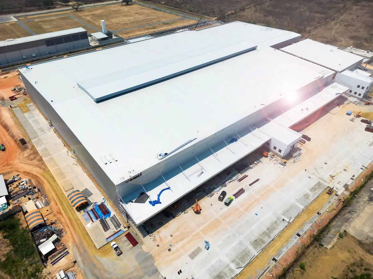 Aerial view of a large, modern industrial warehouse featuring a white commercial roof type, surrounded by construction materials and vehicles on a dirt and concrete site.