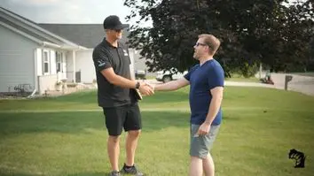 Two men shaking hands outdoors on a grassy lawn. One wears a black cap, shirt, and shorts, while the other wears glasses, a blue shirt, and gray shorts. They stand in front of a suburban house with a tree in the background.