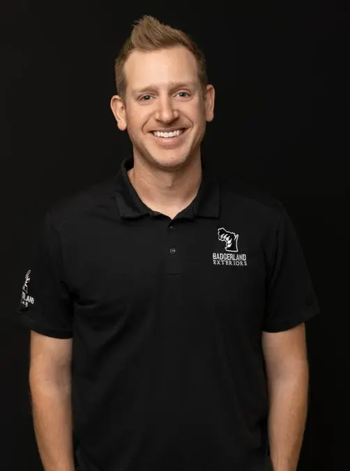 A smiling man with short blond hair wears a black polo shirt with a 